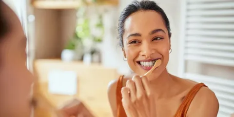 A person smiling while brushing their teeth with a wooden toothbrush, illustrating the action that temporarily softens tooth enamel, making it vulnerable to the acids and pigments in coffee.